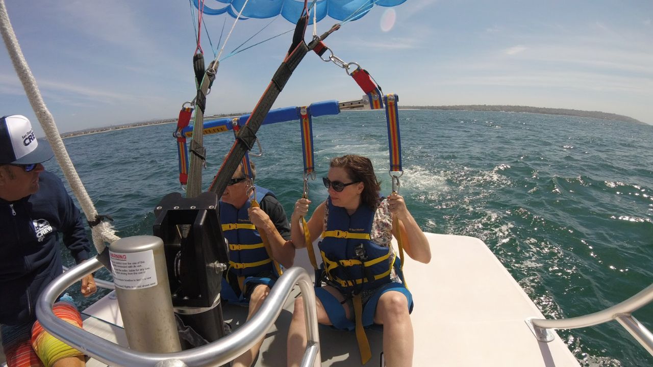 Two people secured in parasailing harnesses preparing for takeoff at the back of a boat with crew nearby