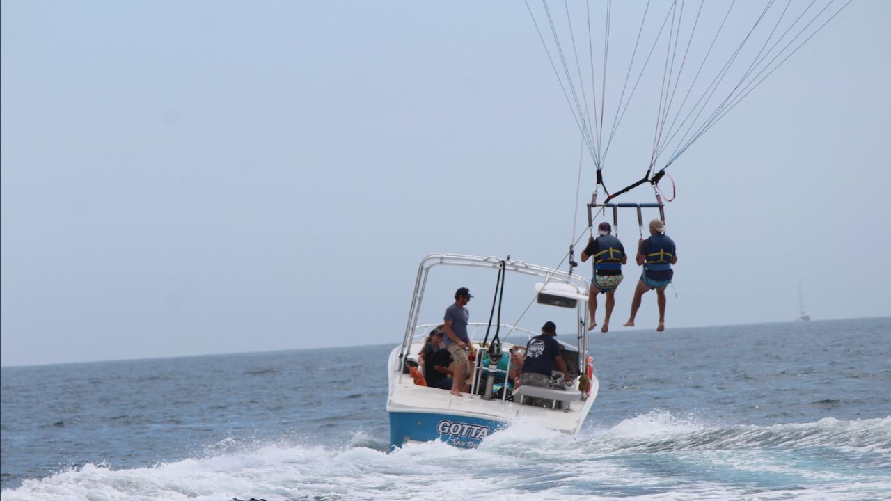 Two riders parasailing behind a speedboat, lifted above ocean waves under a clear sky