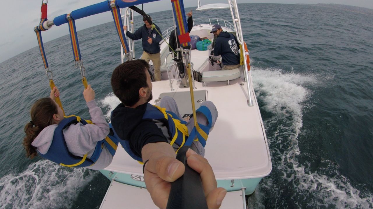 View from boat showing two people harnessed for parasailing with crew onboard.