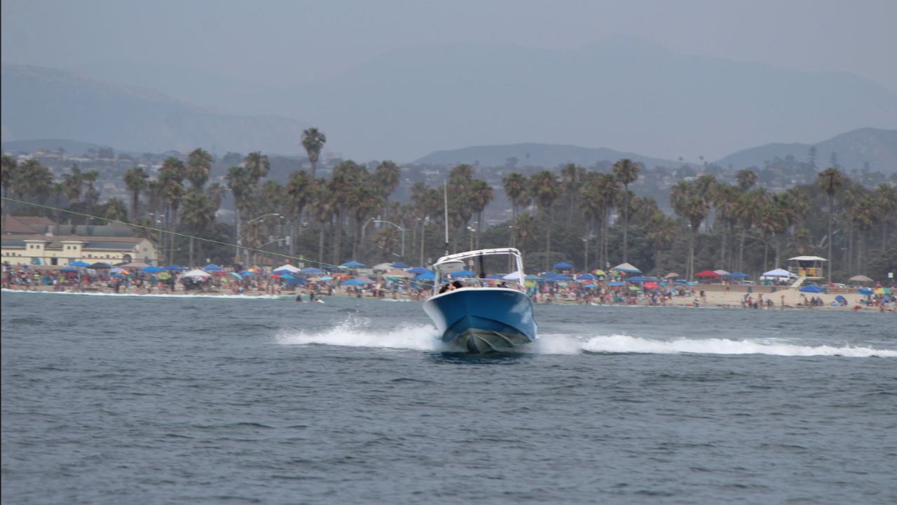 Speedboat towing a parasail line across the water near a busy beach with palm trees and mountain views in the background