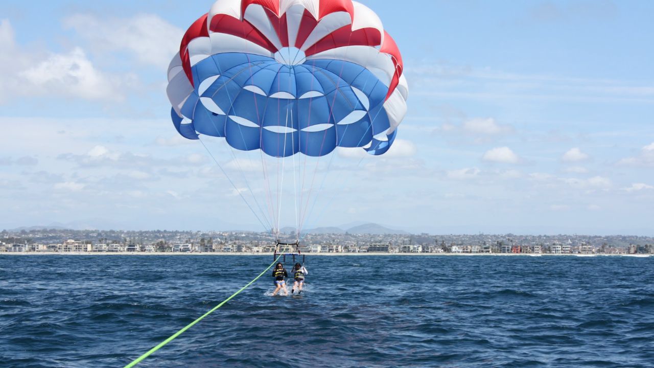 Two people parasailing above Mission Bay with the San Diego coastline in the background.