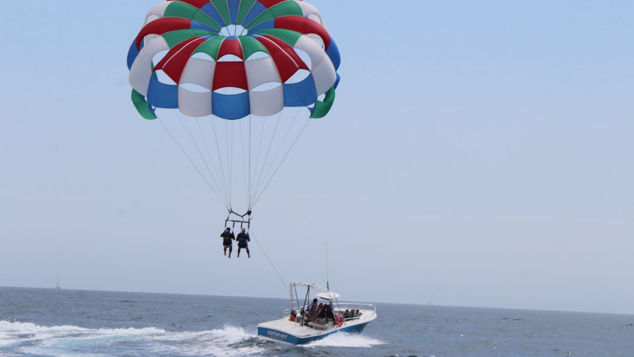 Two guests parasailing high above the ocean behind a boat beneath a colorful parachute.