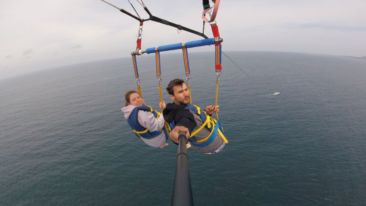 Two people tandem parasailing high above the ocean while wearing life jackets and secure harnesses in San Diego.