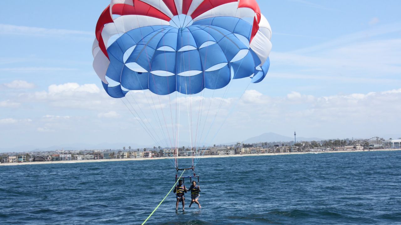 Two guests enjoying tandem parasailing beneath a colorful parachute with the San Diego coastline in the background.