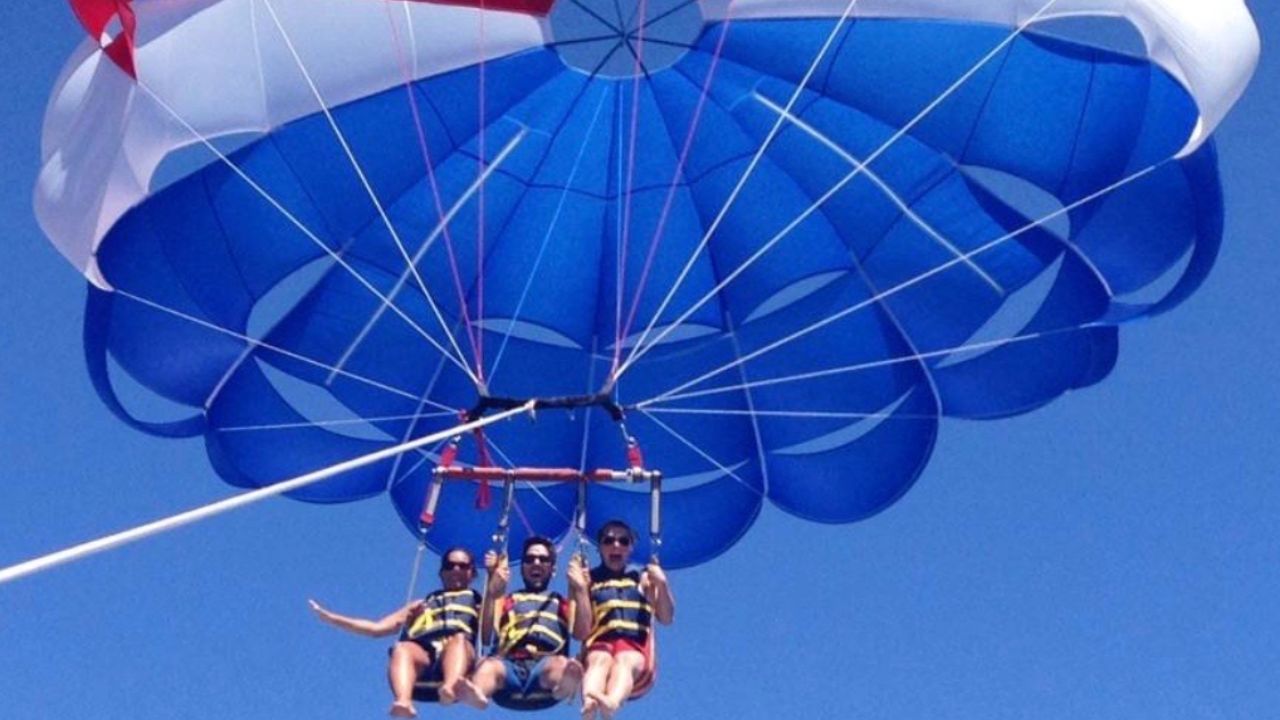 Three people enjoying a tandem parasailing flight beneath a colorful parachute over the Pacific Ocean.