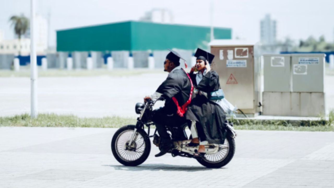 Two graduates in caps and gowns ride motorcycle outdoors celebrating.